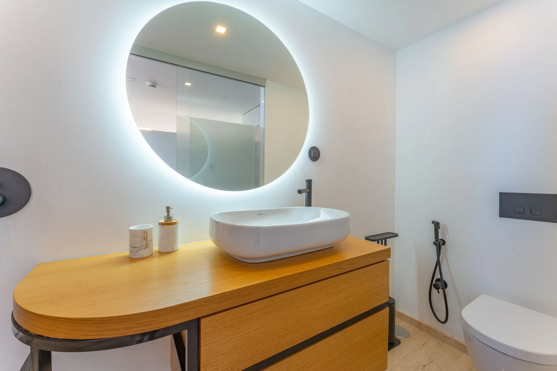 A modern bathroom with a vessel sink, illuminated round mirror, and shower.