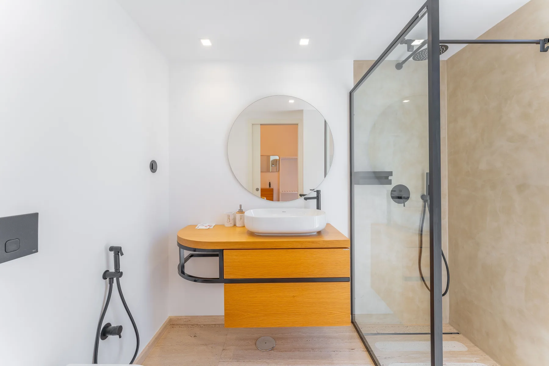 A bright bathroom with a wood vanity, round mirror, and glass shower.