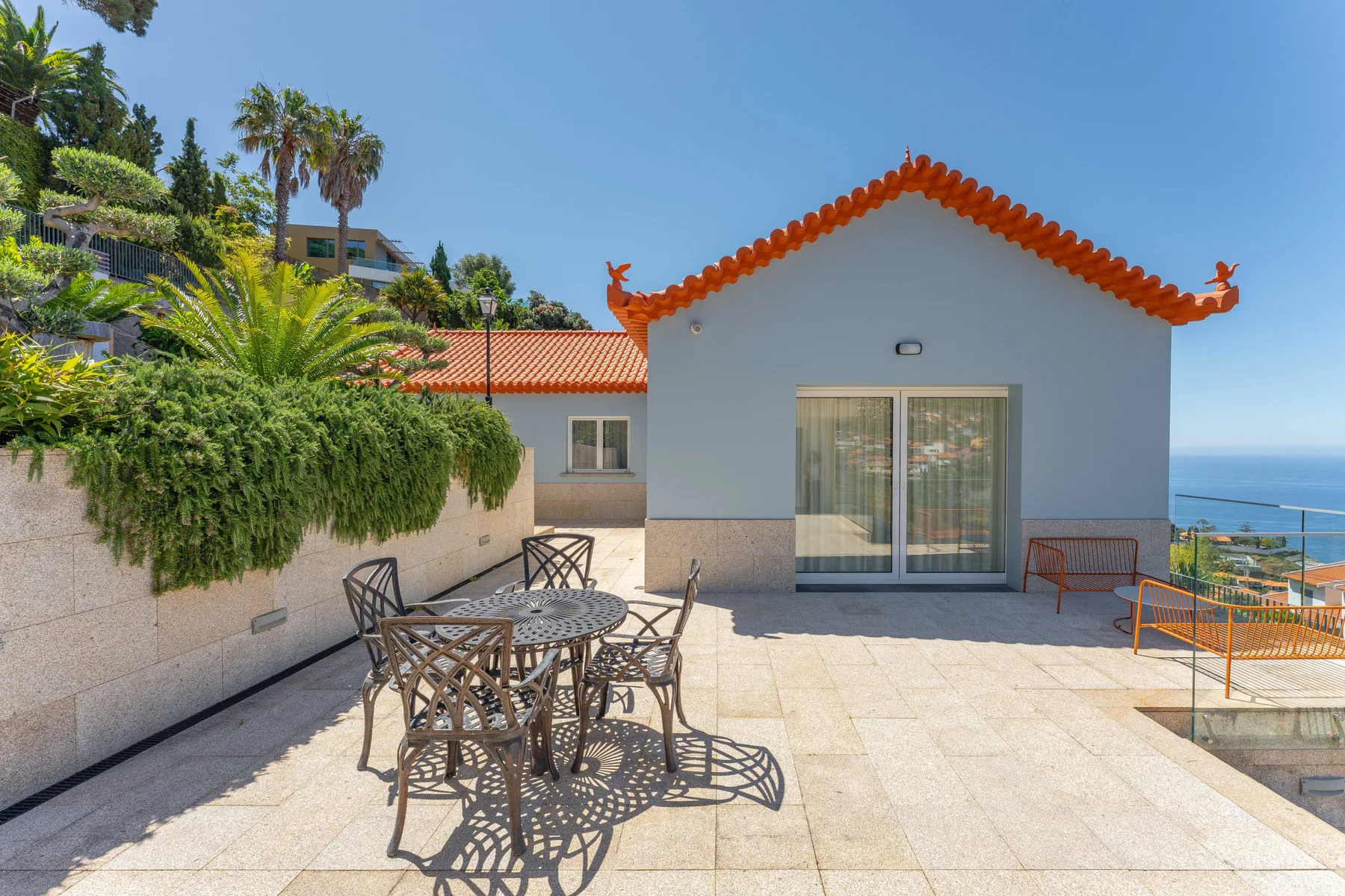 A paved outdoor terrace has a metal dining set beside a white house with an orange-tiled roof.