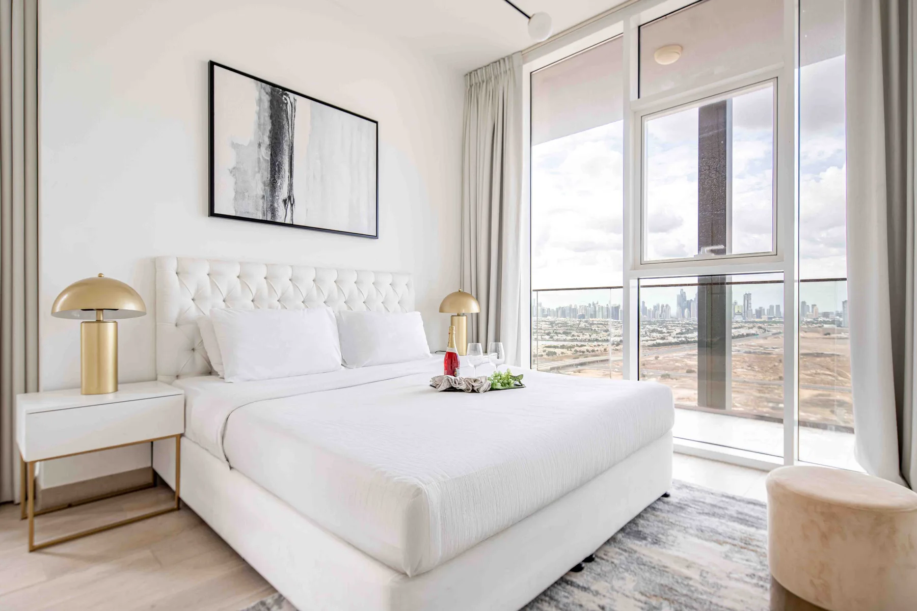 A bright bedroom with a white bed beside large windows overlooking the city.
