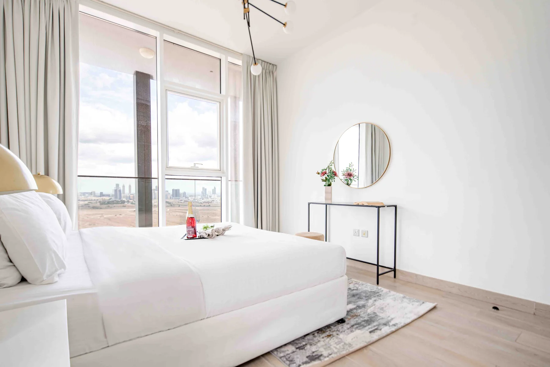 A bright bedroom with a white bed, tall windows, and a round wall mirror.