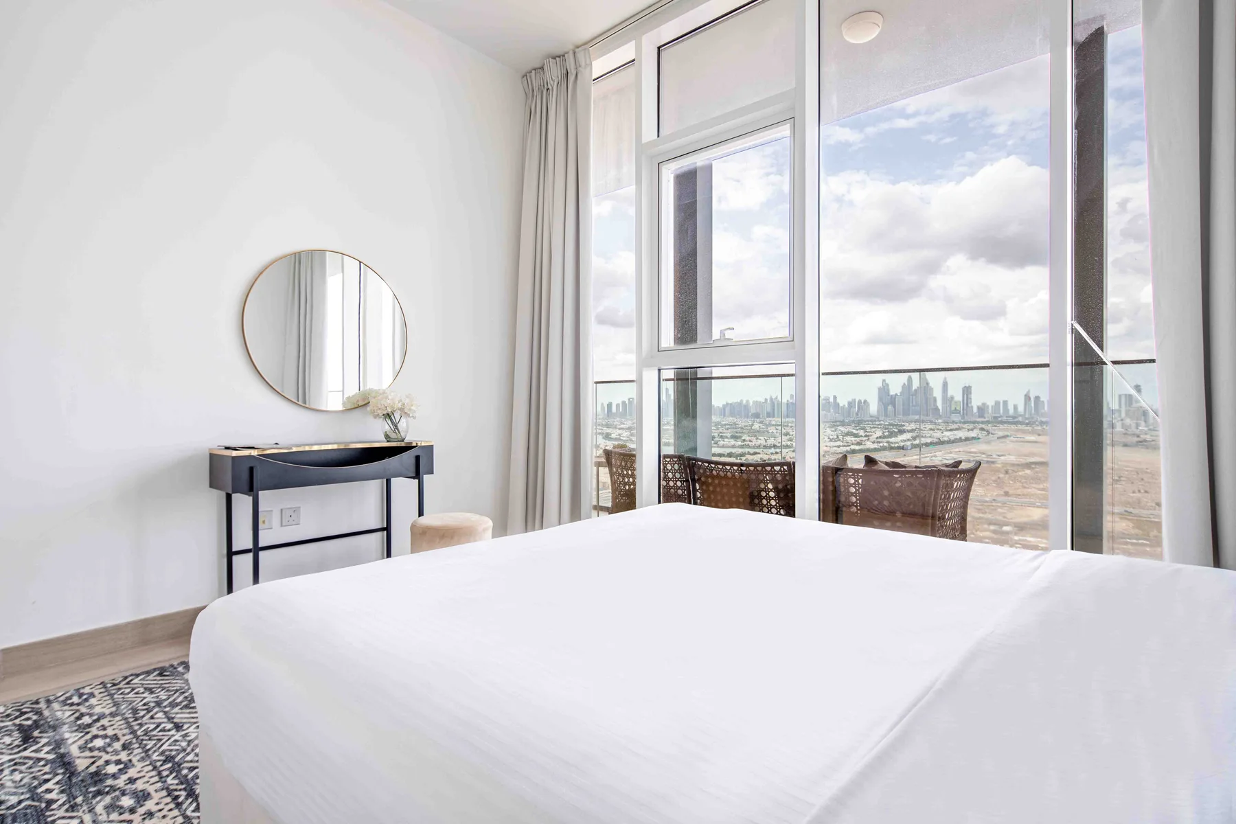 A bedroom with a neatly made bed beside large windows overlooking a city skyline.