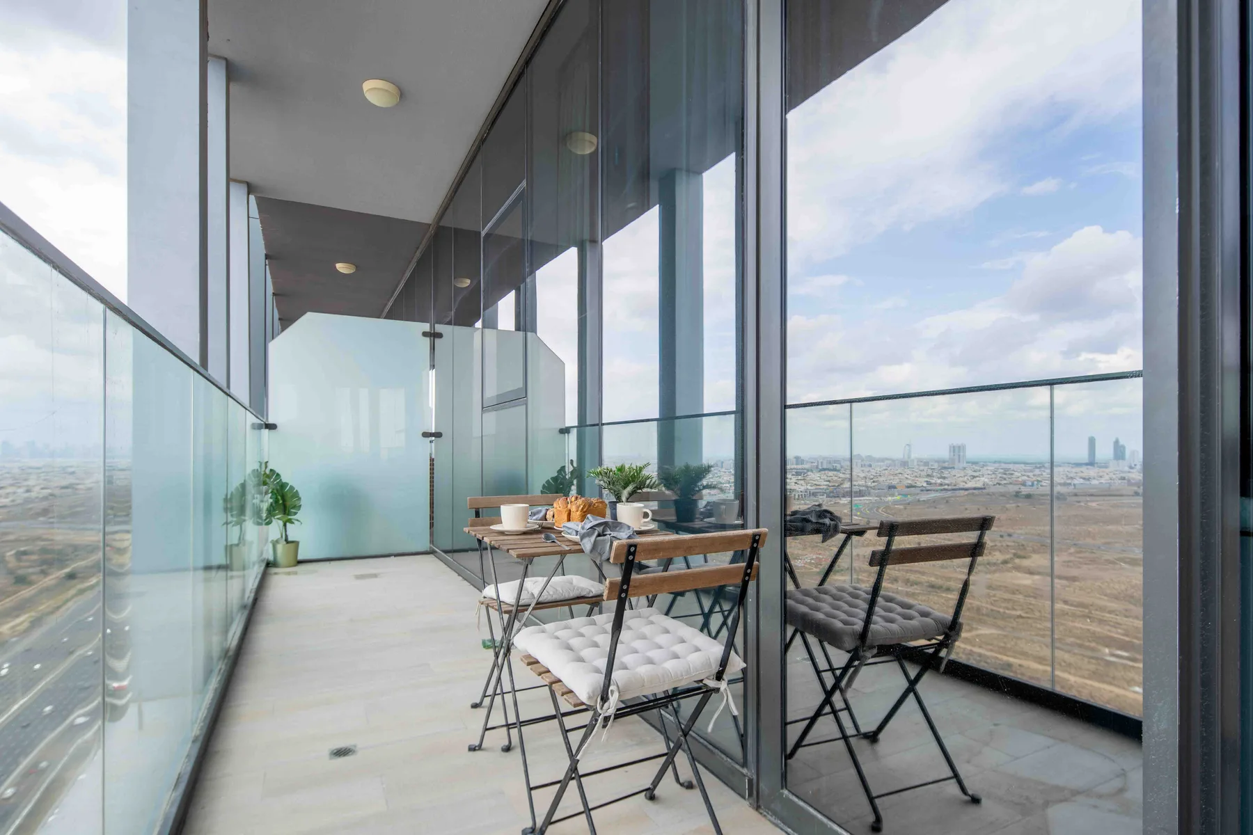 A glass-walled balcony with a small dining table and folding chairs overlooks the city.