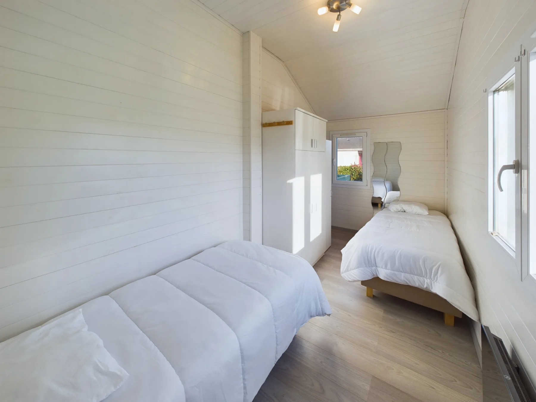 A bright bedroom with two beds and white paneled walls.