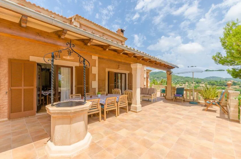 A tiled outdoor terrace beside a house has seating and a small fountain.