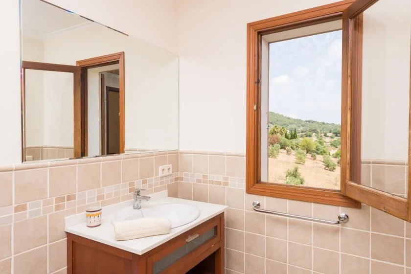 A tiled bathroom with a sink and an open window overlooking a green landscape.