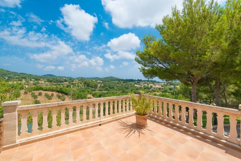 A sunlit terrace overlooks green hills, with a potted plant beside the railing.