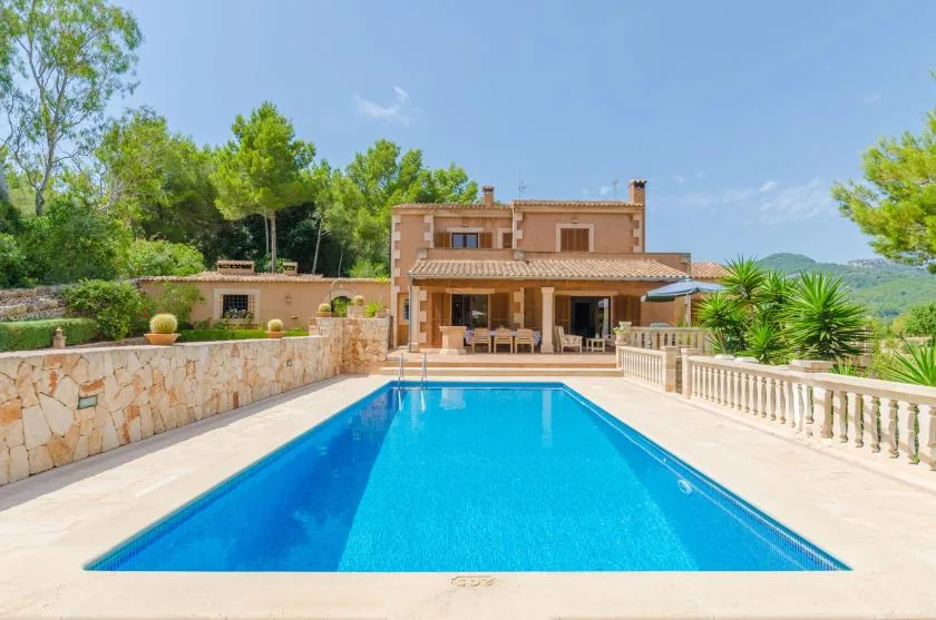 A swimming pool faces a two-story house, with trees and greenery around the patio.