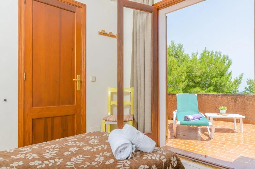 A bedroom opens onto a sunlit terrace with outdoor seating and green trees.