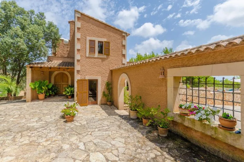 A sunlit stone courtyard sits beside a warm-toned building with arched openings and potted plants.