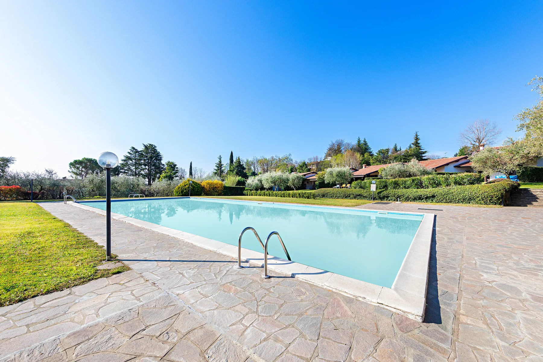An outdoor swimming pool is surrounded by paved areas and landscaped greenery.