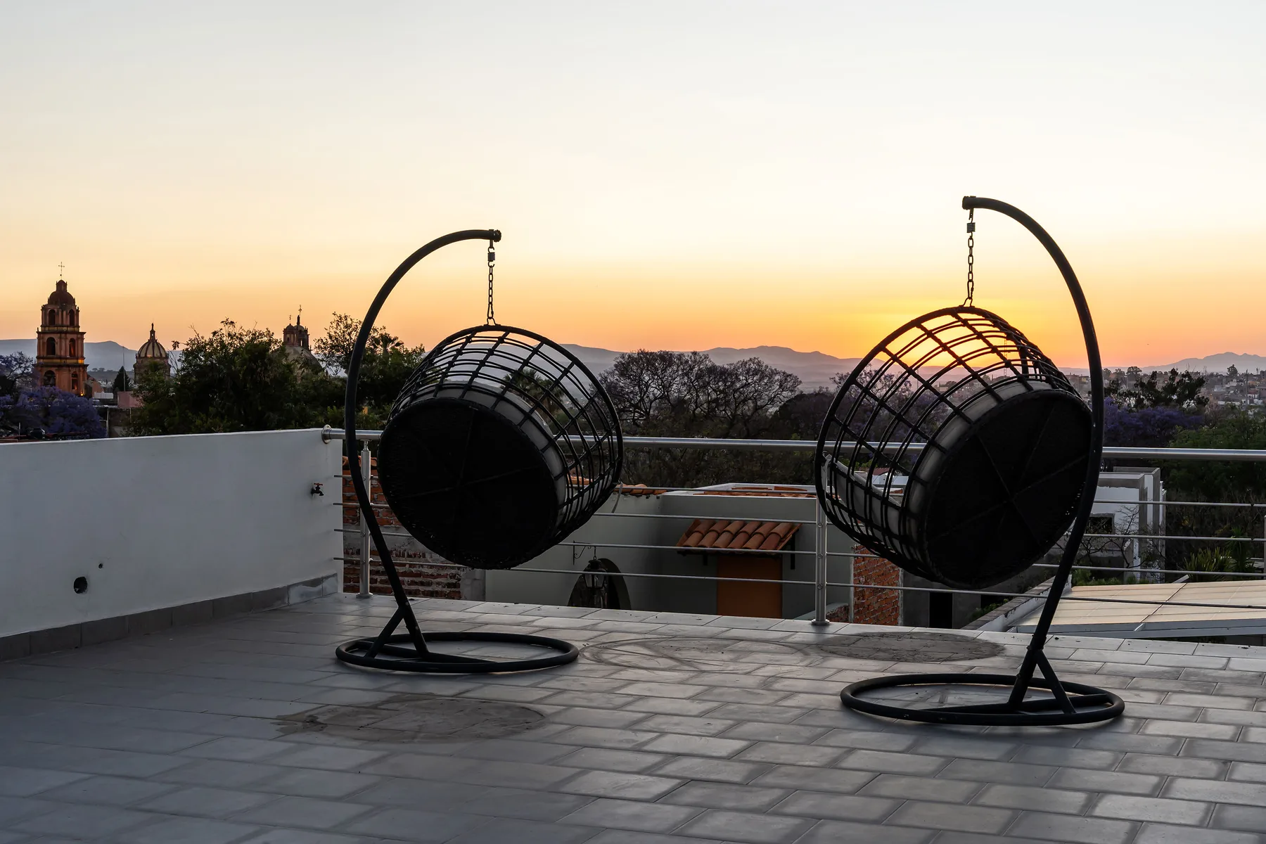 A rooftop terrace with two suspended seats overlooking a city at sunset.