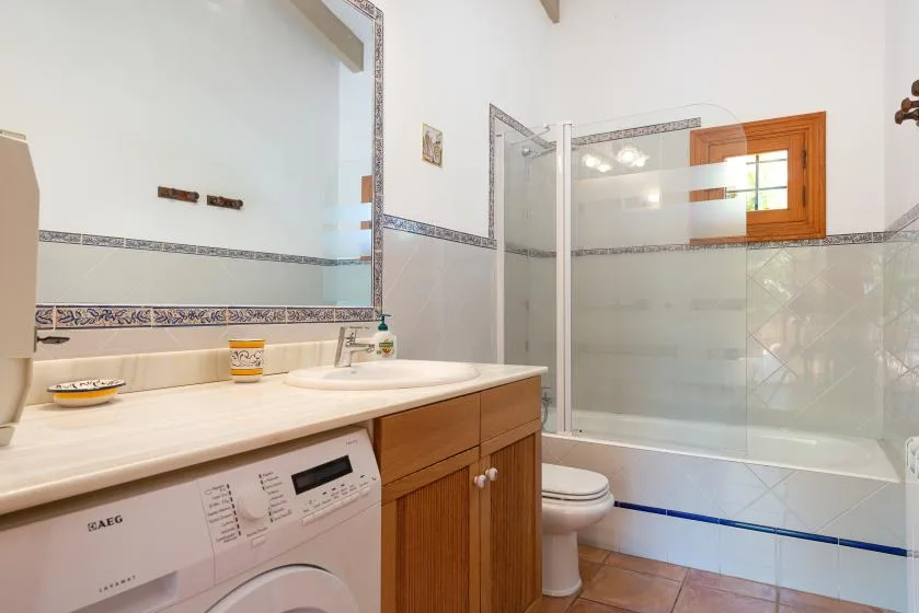 A bathroom with a sink, washing machine, toilet, and bathtub behind a glass screen.