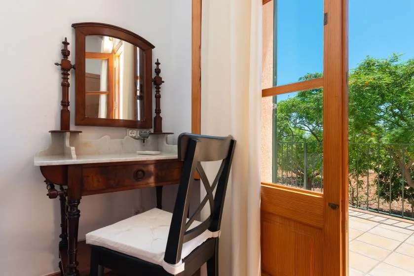 A wooden vanity and chair sit beside an open glass door overlooking greenery.