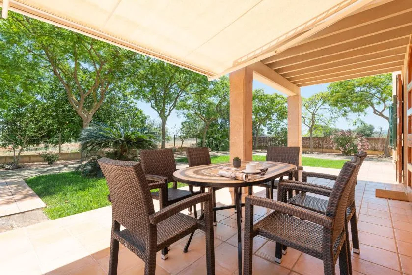 A covered outdoor patio has a dining table and woven seating beside a garden.