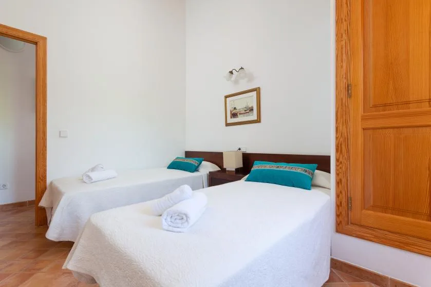A bright bedroom with two beds dressed in white linens and teal cushions.