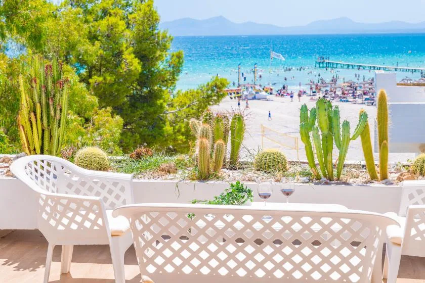 Una terraza junto al mar con asientos blancos y cactus frente a una playa y aguas azules.