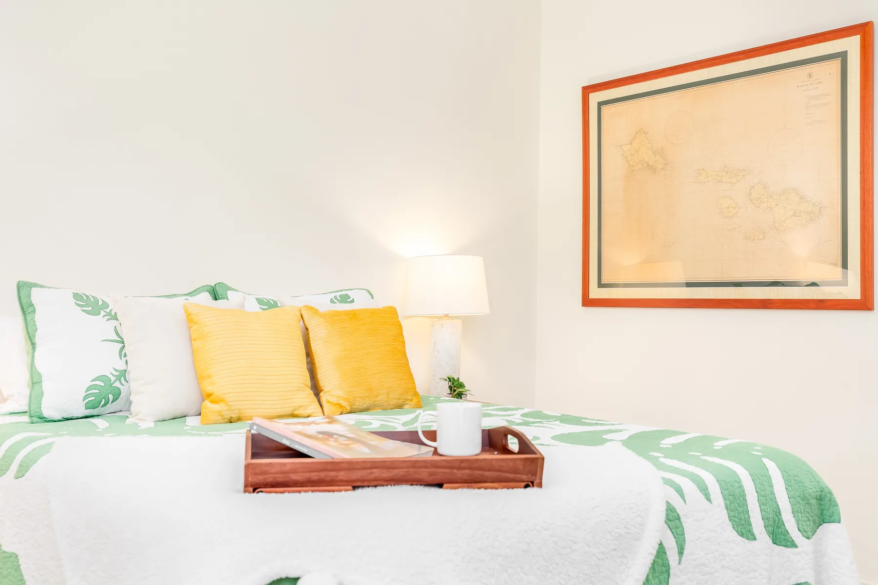 Una cama con ropa de cama verde y blanca, almohadas amarillas y una bandeja está bajo un cuadro enmarcado.