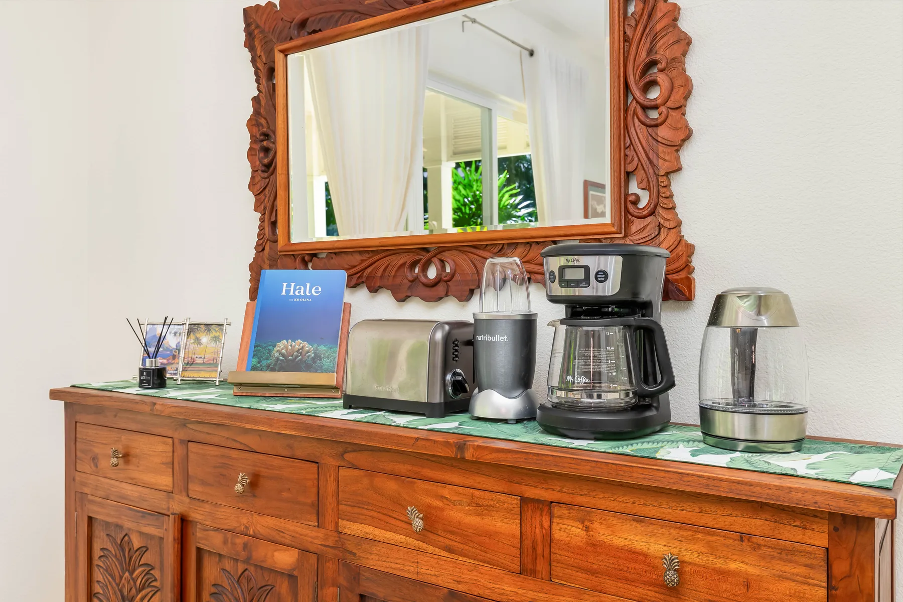 Un aparador de madera muestra una cafetera, un hervidor y un espejo enmarcado.