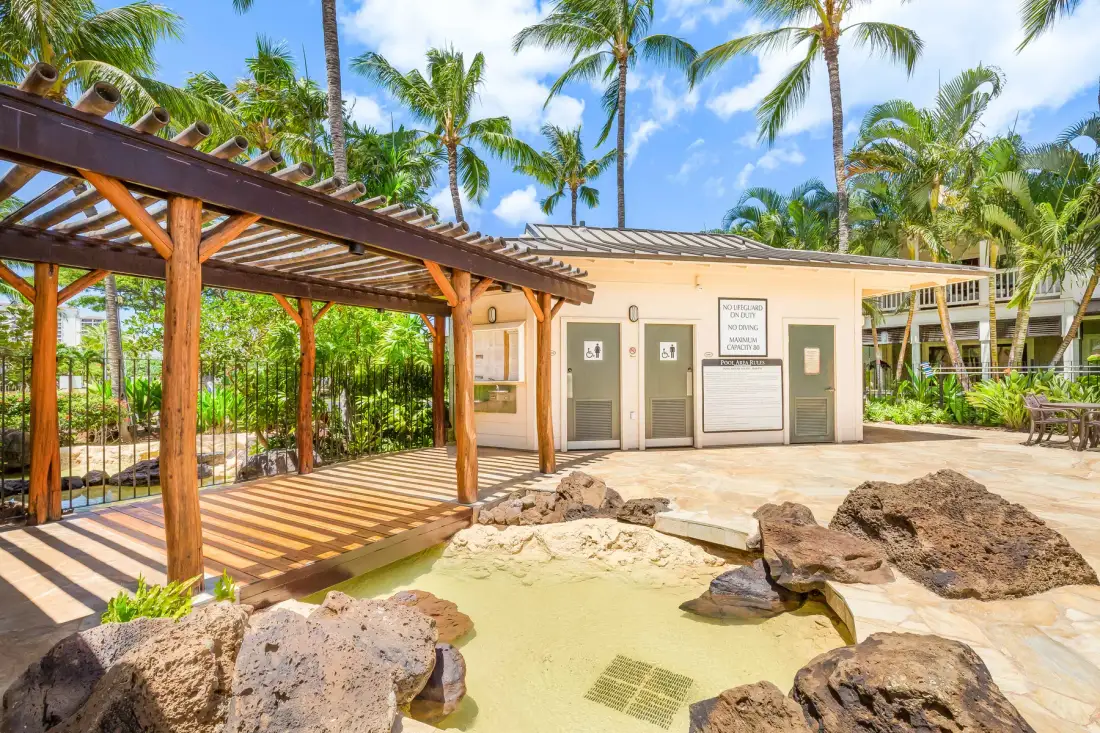 Una piscina exterior poco profunda está junto a una pérgola de madera, rodeada de palmeras.