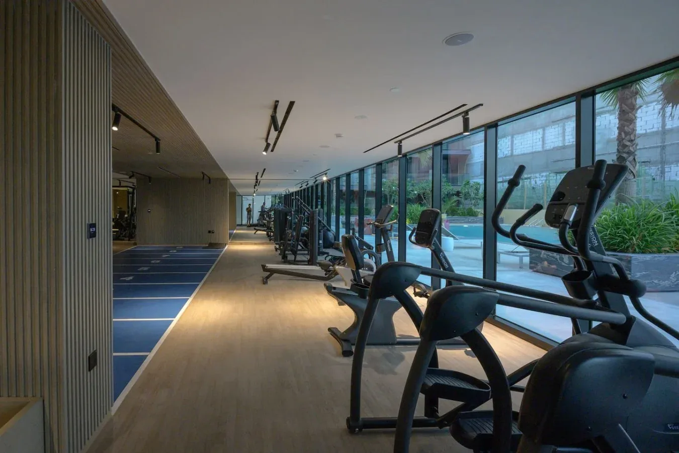 A gym with exercise machines beside large windows overlooking an outdoor pool.