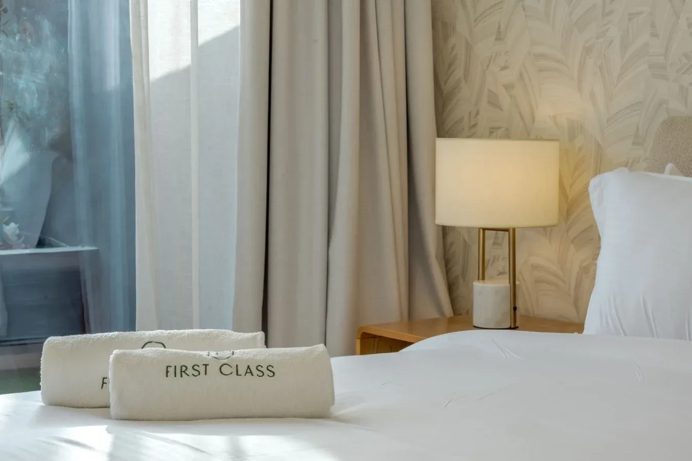 A bedroom with a neatly made bed, bedside lamp, and neutral patterned wallpaper.