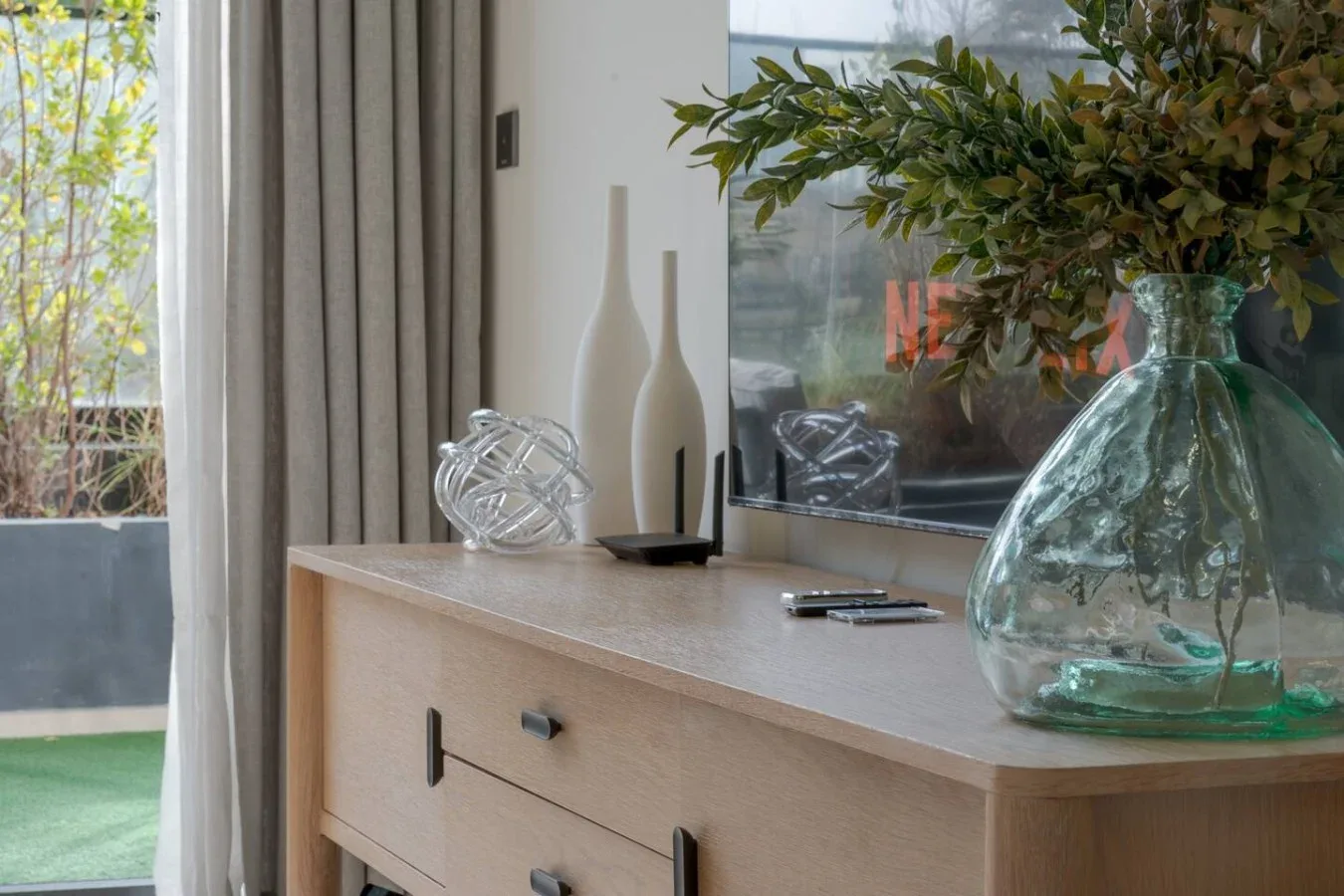 A bright interior console table displays glass vases beside a large window.