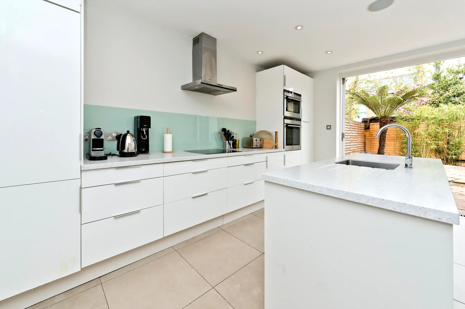 A bright modern kitchen with white cabinetry, pale green backsplash, and a large island.