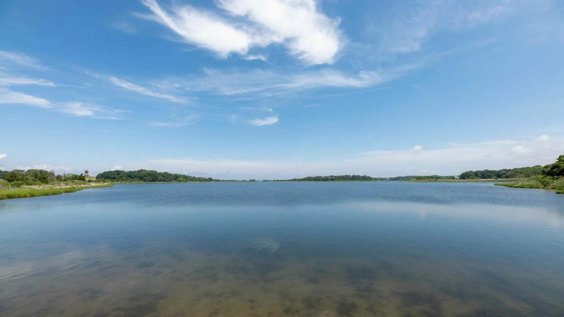 Un amplio lago se extiende bajo un cielo azul, con orillas verdes bajas a lo lejos.