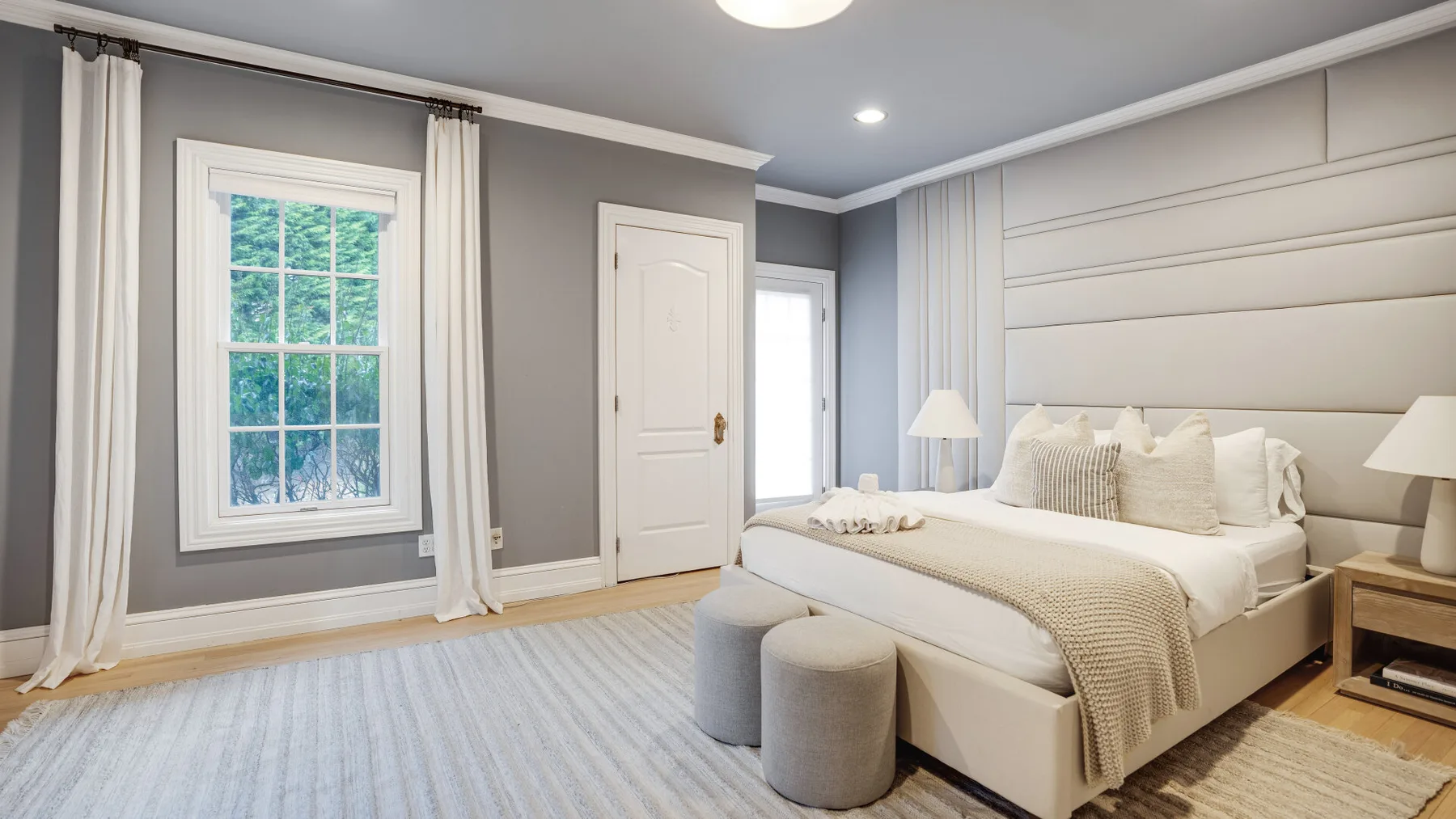 Un dormitorio luminoso con una cama grande, ropa de cama clara y ventanas altas.