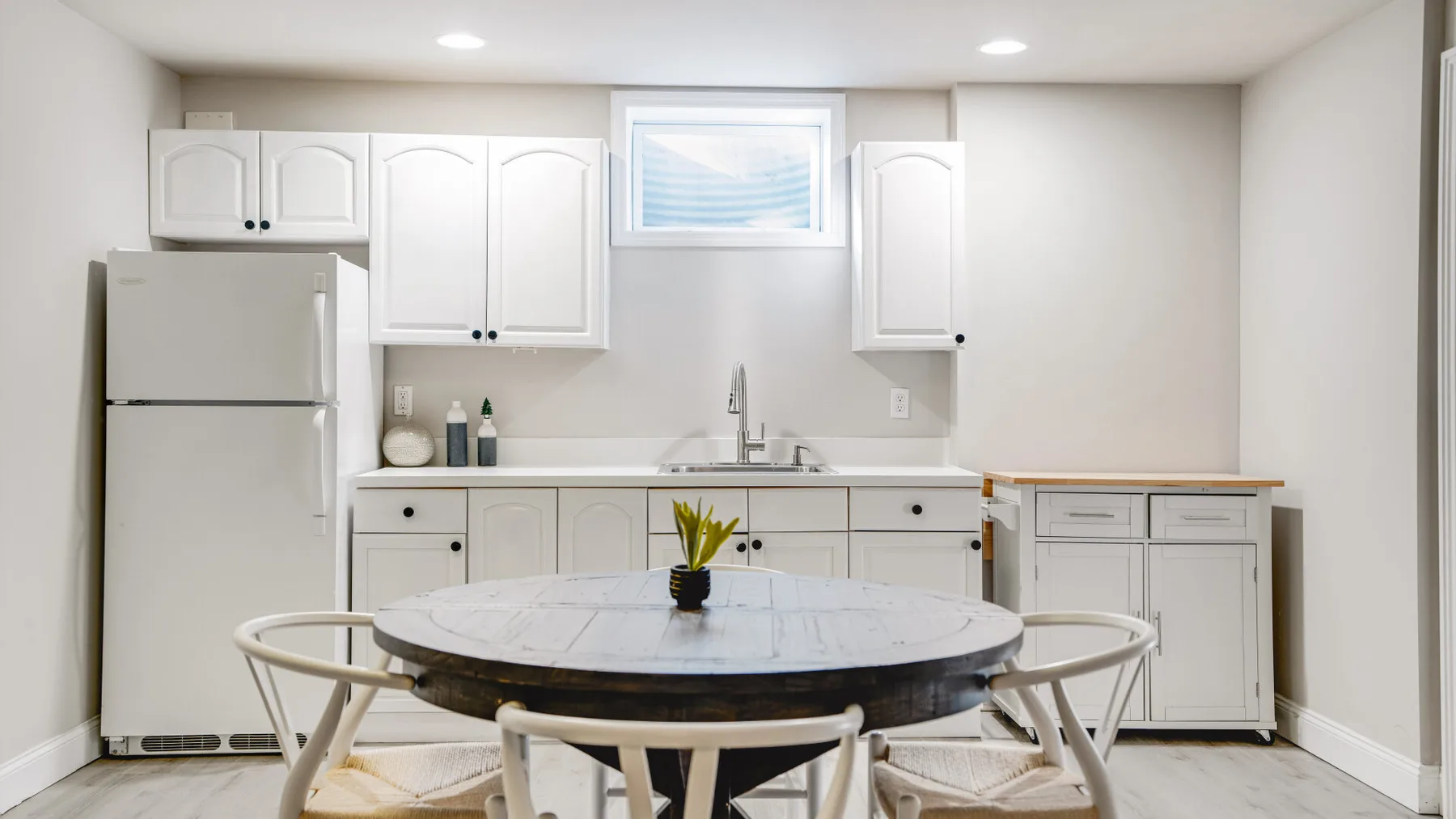 Una cocina compacta con gabinetes blancos, un refrigerador y una mesa de comedor.