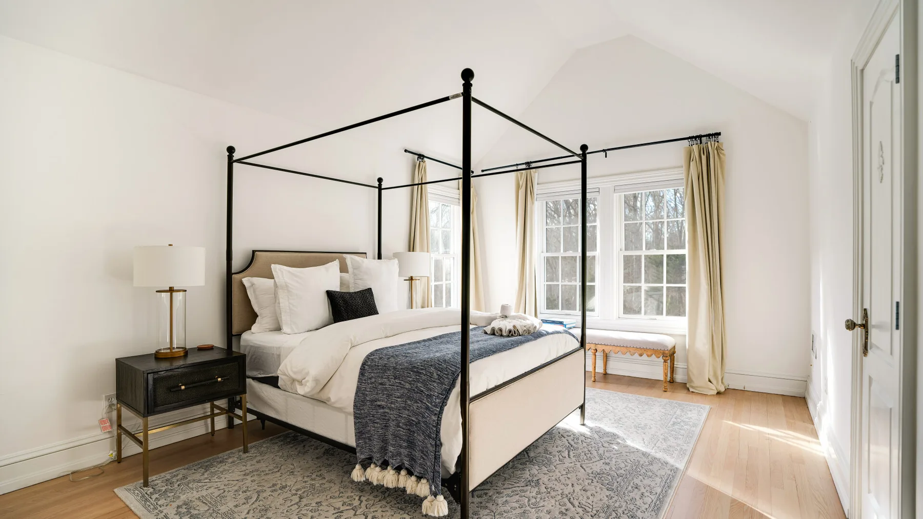 Un dormitorio luminoso con cama con dosel, ventanas grandes y una alfombra estampada.