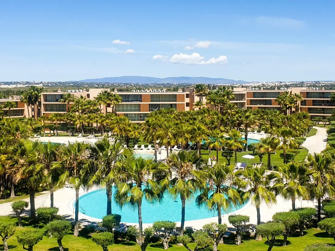 Vista de un complejo ajardinado con piscina, palmeras y edificios bajos.