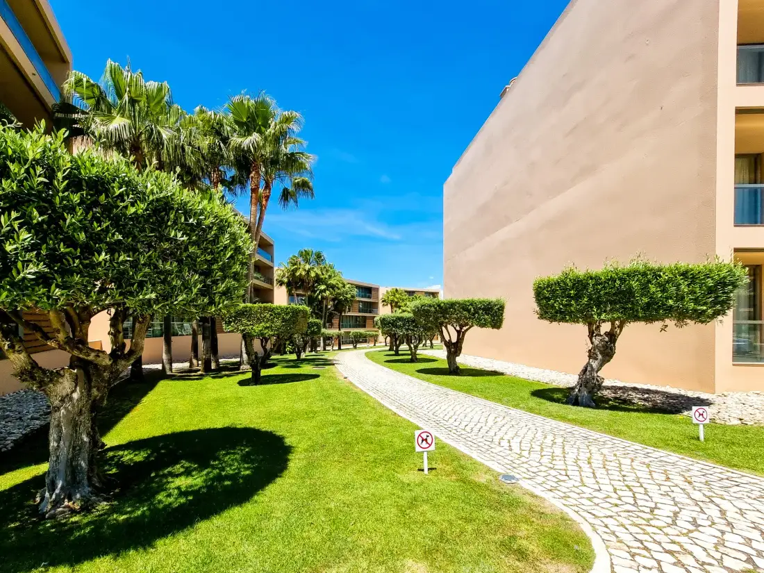 Un patio ajardinado de apartamentos tiene un sendero curvo de adoquines entre césped y árboles podados.