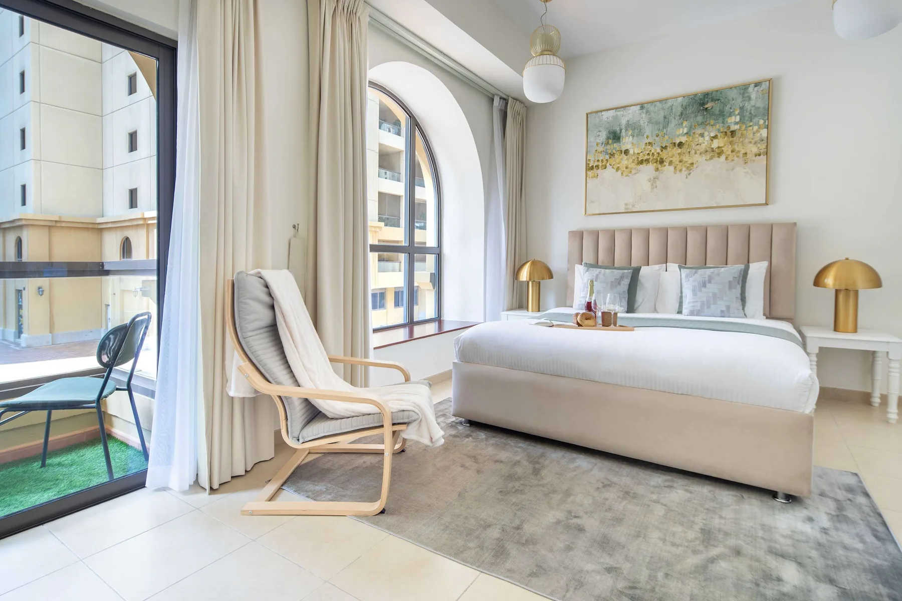 Un dormitorio luminoso con una cama, un sillón acolchado y ventanas altas con arco.