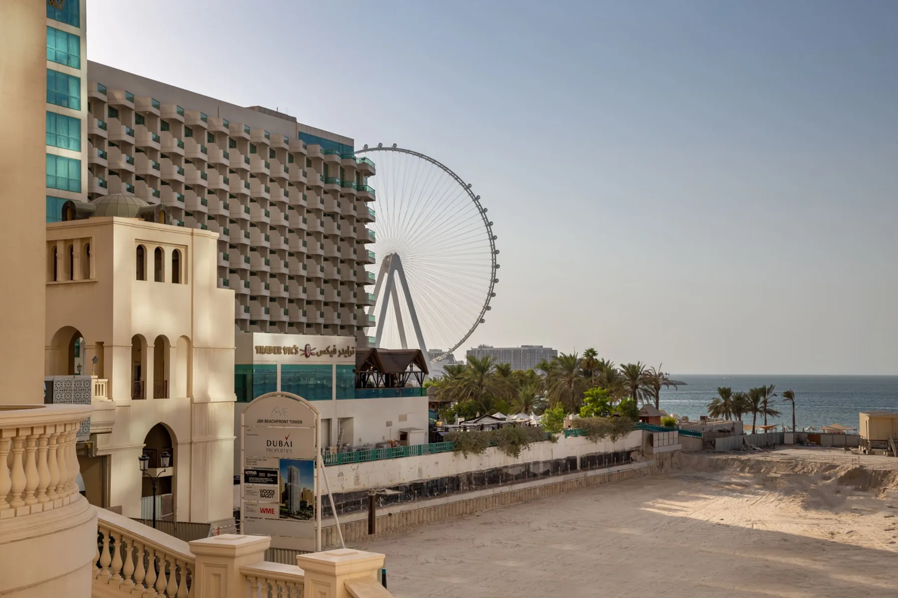 Una vista frente al mar muestra una gran noria junto a edificios hoteleros y palmeras.