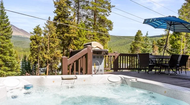 An outdoor hot tub sits beside a wooden deck with seating and a mountain view.