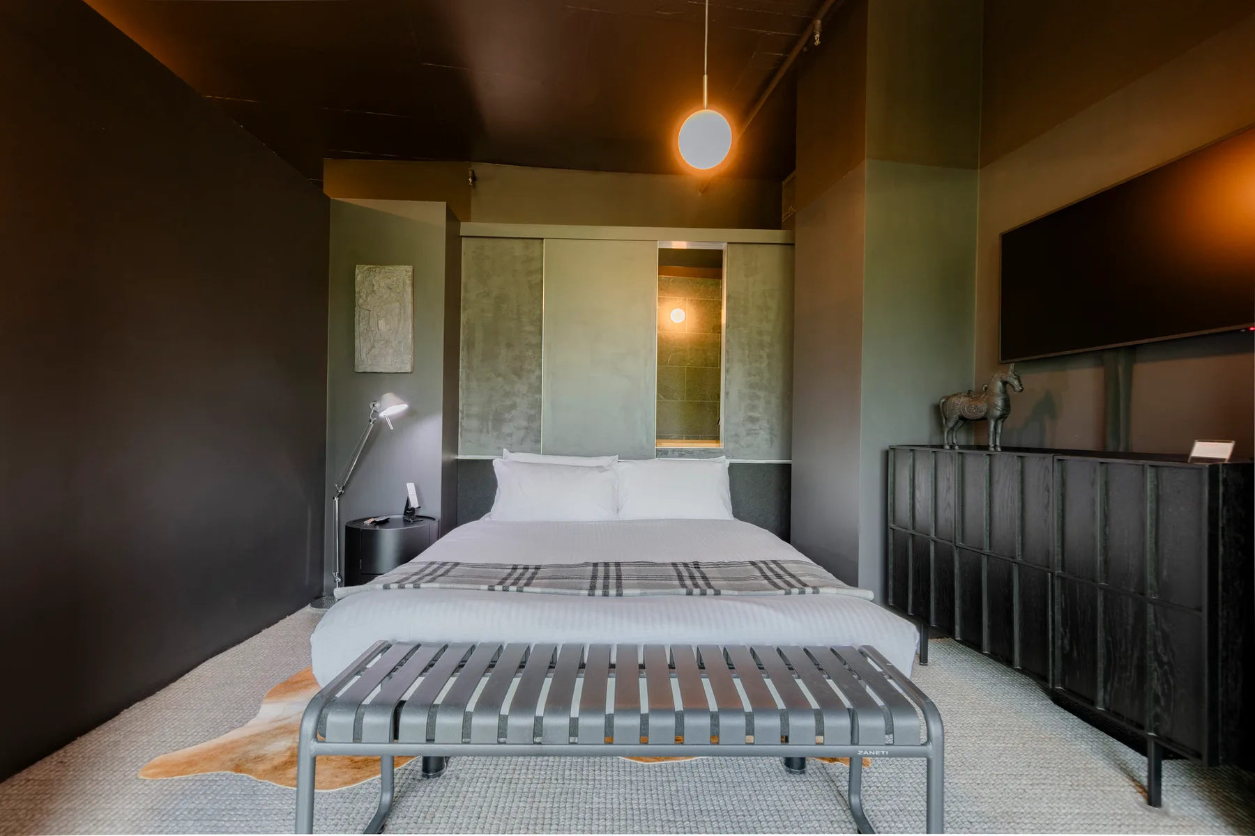Un dormitorio con una cama, un banco metálico y paredes oscuras.
