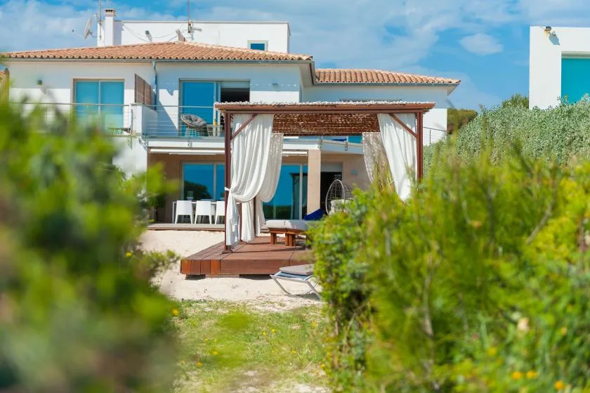 Una casa blanca con una pérgola de madera con cortinas y vegetación en primer plano.