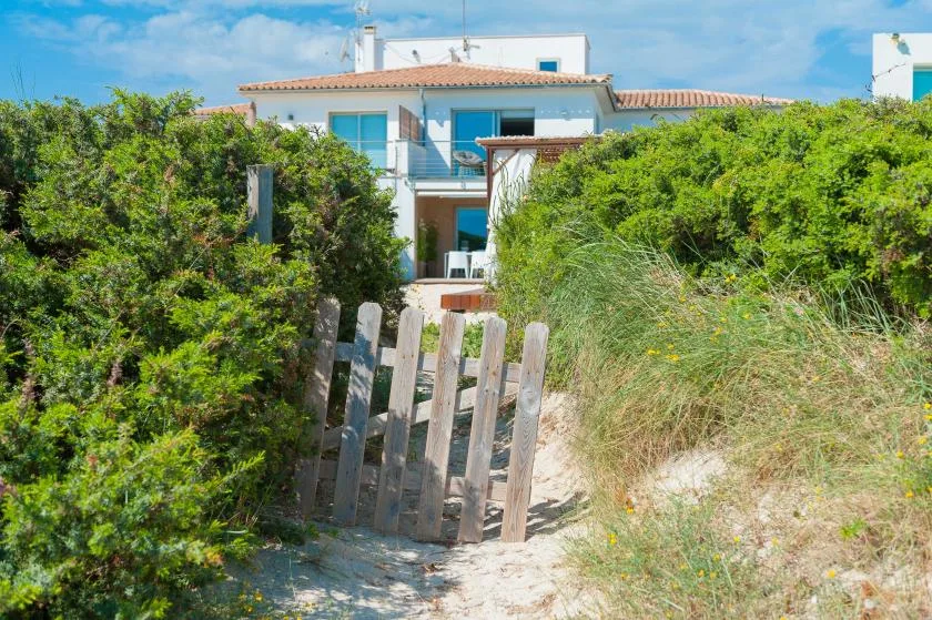 Un sendero de arena atraviesa la vegetación costera hacia una casa blanca.