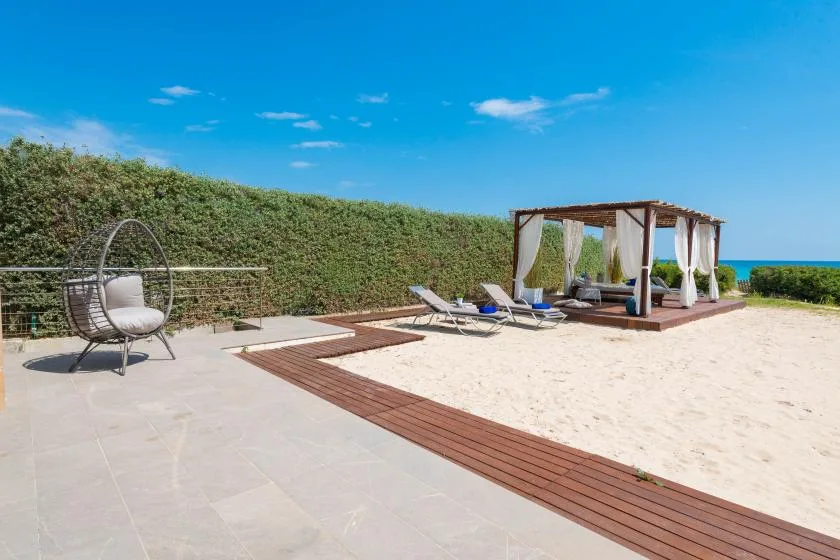 Un patio soleado da a una playa de arena, con tumbonas y una zona de estar con sombra.