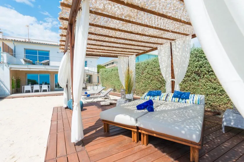 Una zona de estar al aire libre bajo una pérgola, con asientos acolchados junto a una piscina.
