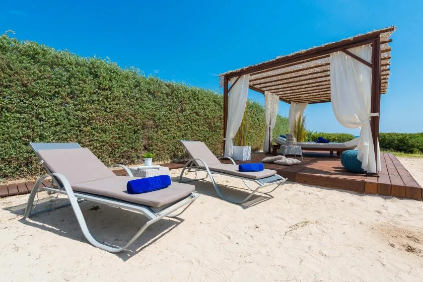 Una zona de descanso exterior sobre arena, con asientos reclinables junto a una pérgola de madera con cortinas.