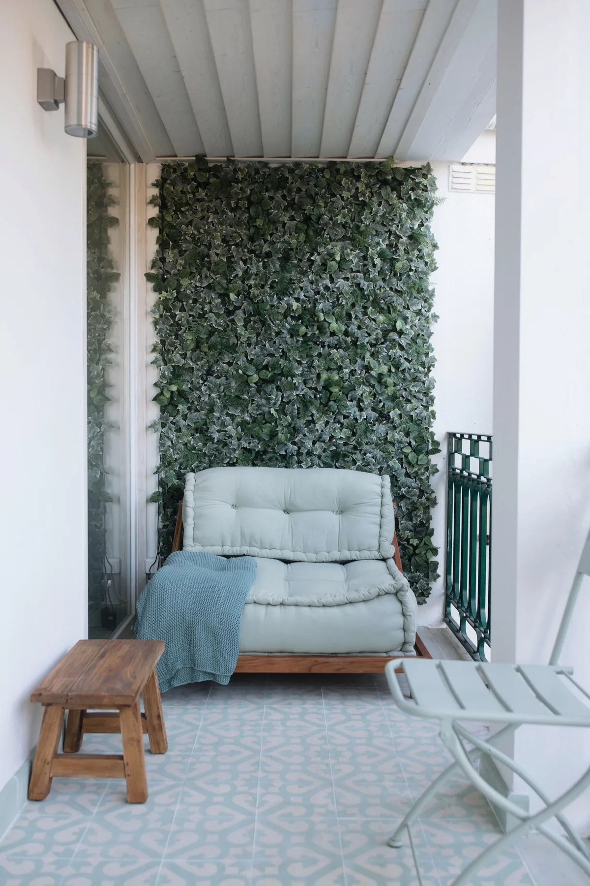 A covered balcony seating area features a cushioned seat beside a leafy wall.