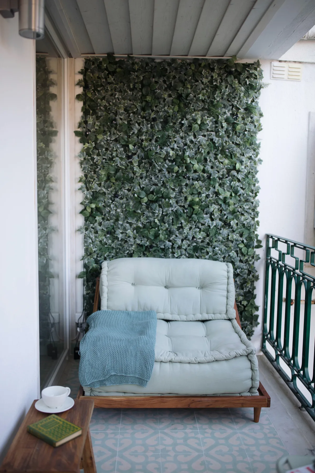 A covered balcony has cushioned seating beside a leafy wall.