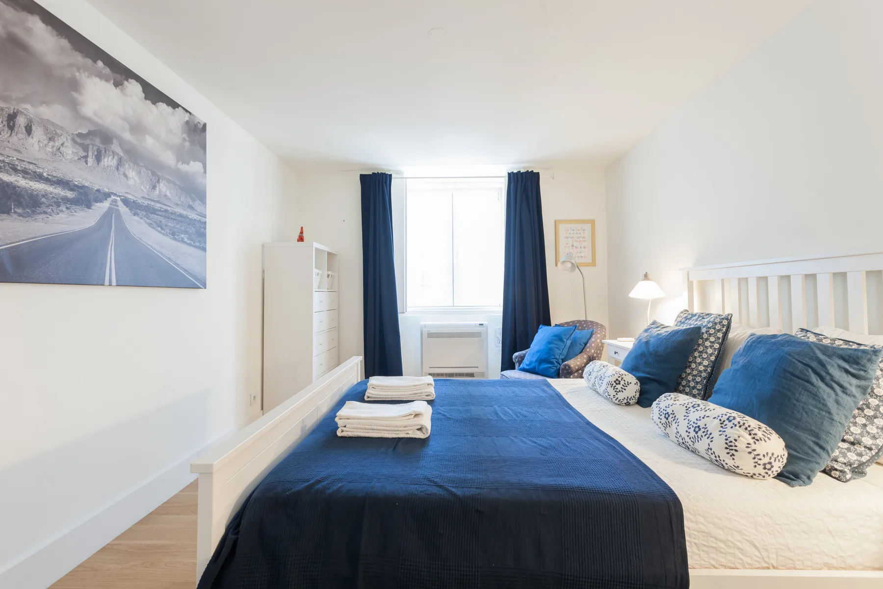 A bright bedroom has a bed with blue bedding and a window with dark curtains.