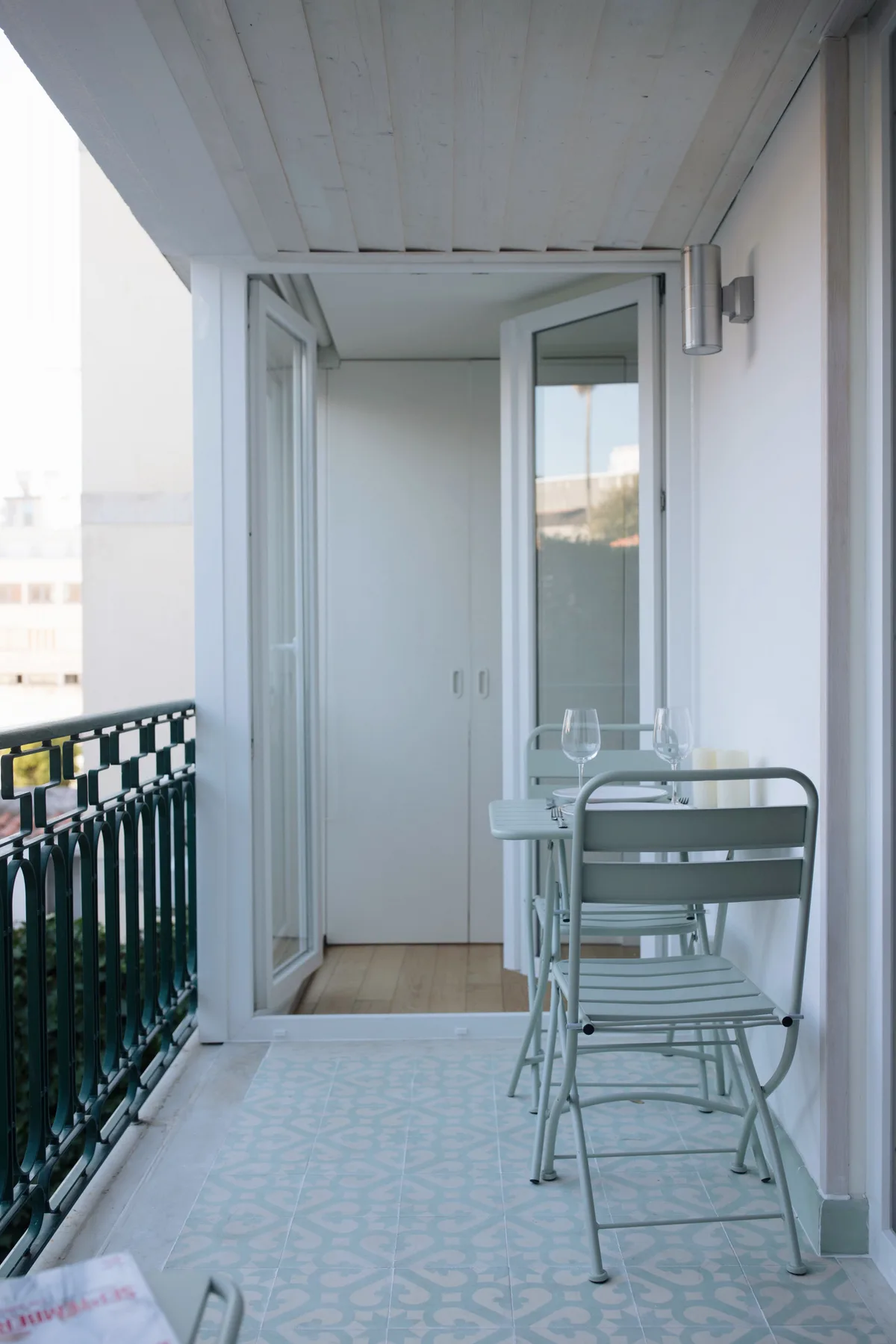 A narrow balcony with patterned floor tiles and metal seating beside open doors.