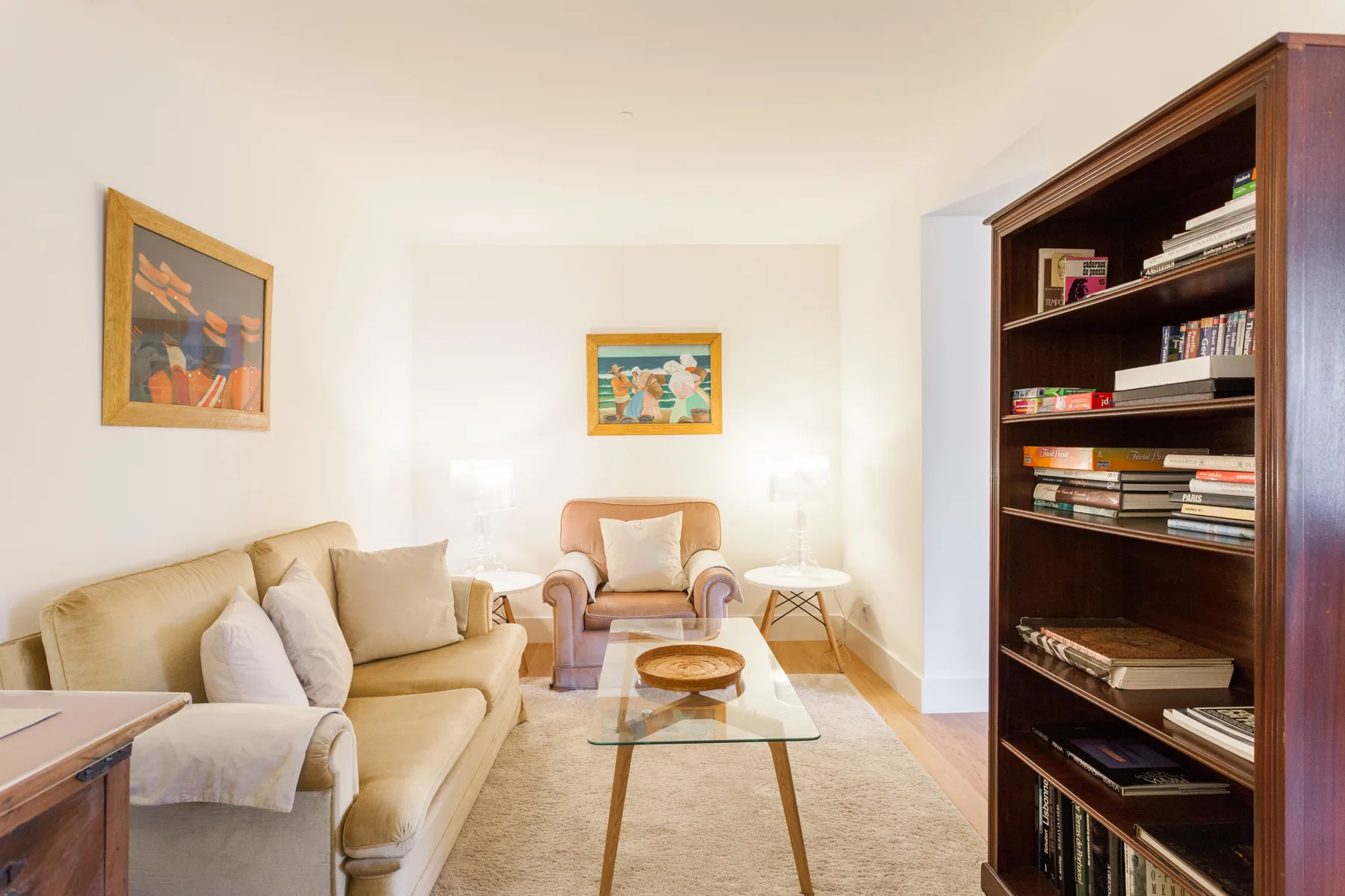 A bright living room with beige seating, a glass coffee table, and a tall bookcase.
