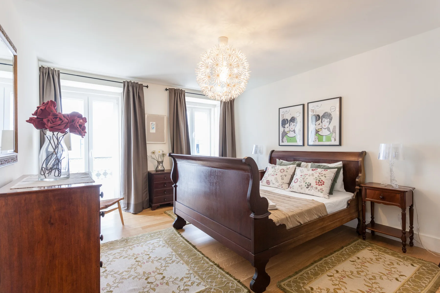 A bright bedroom with a wooden bed, patterned bedding, and tall windows.