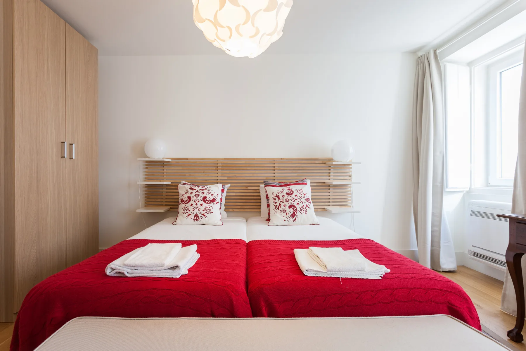 A bright bedroom with two beds dressed in red and a large window.
